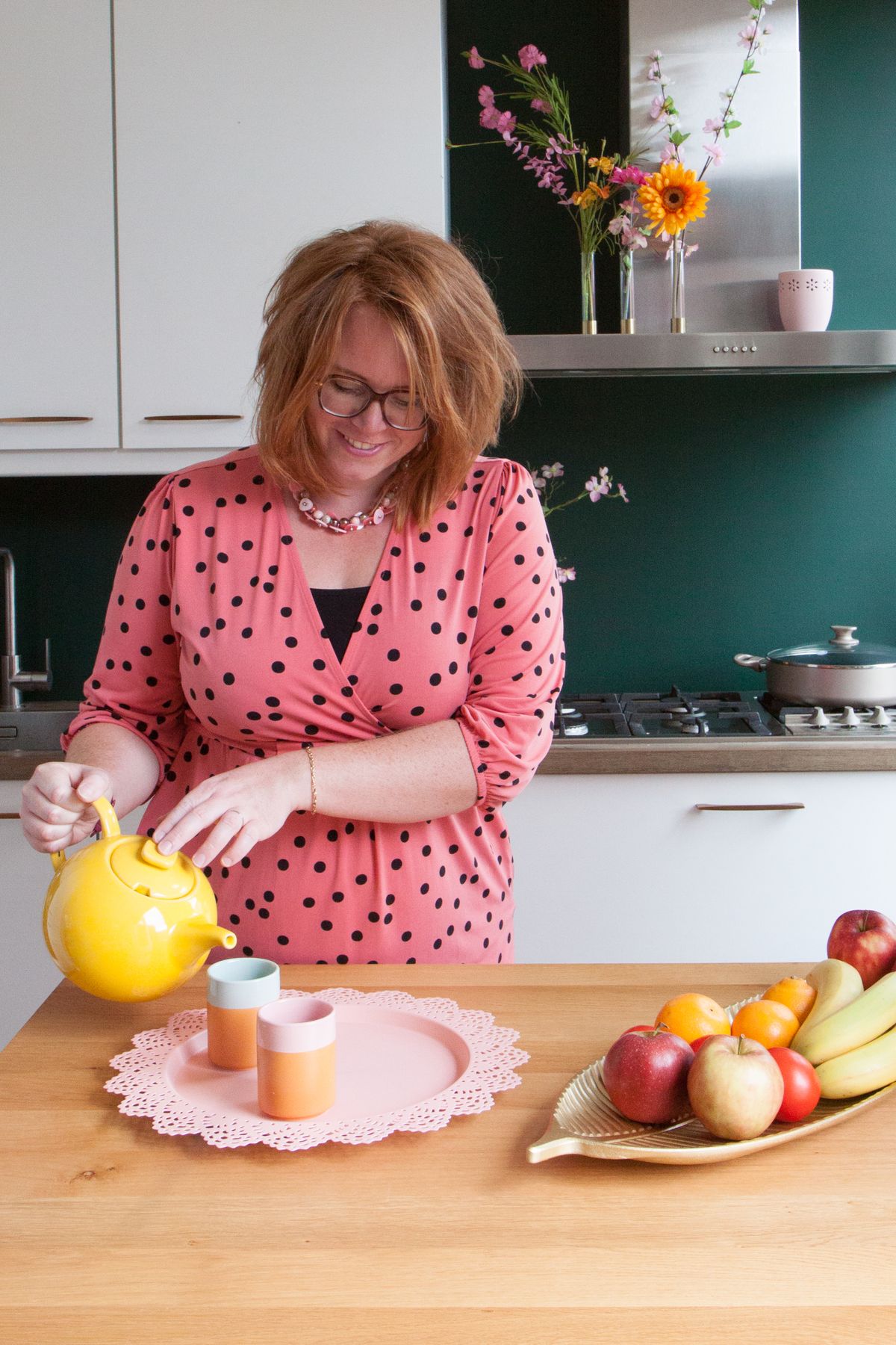 Roodharige dame schenkt thee in, roze stippeljurk in de keuken.