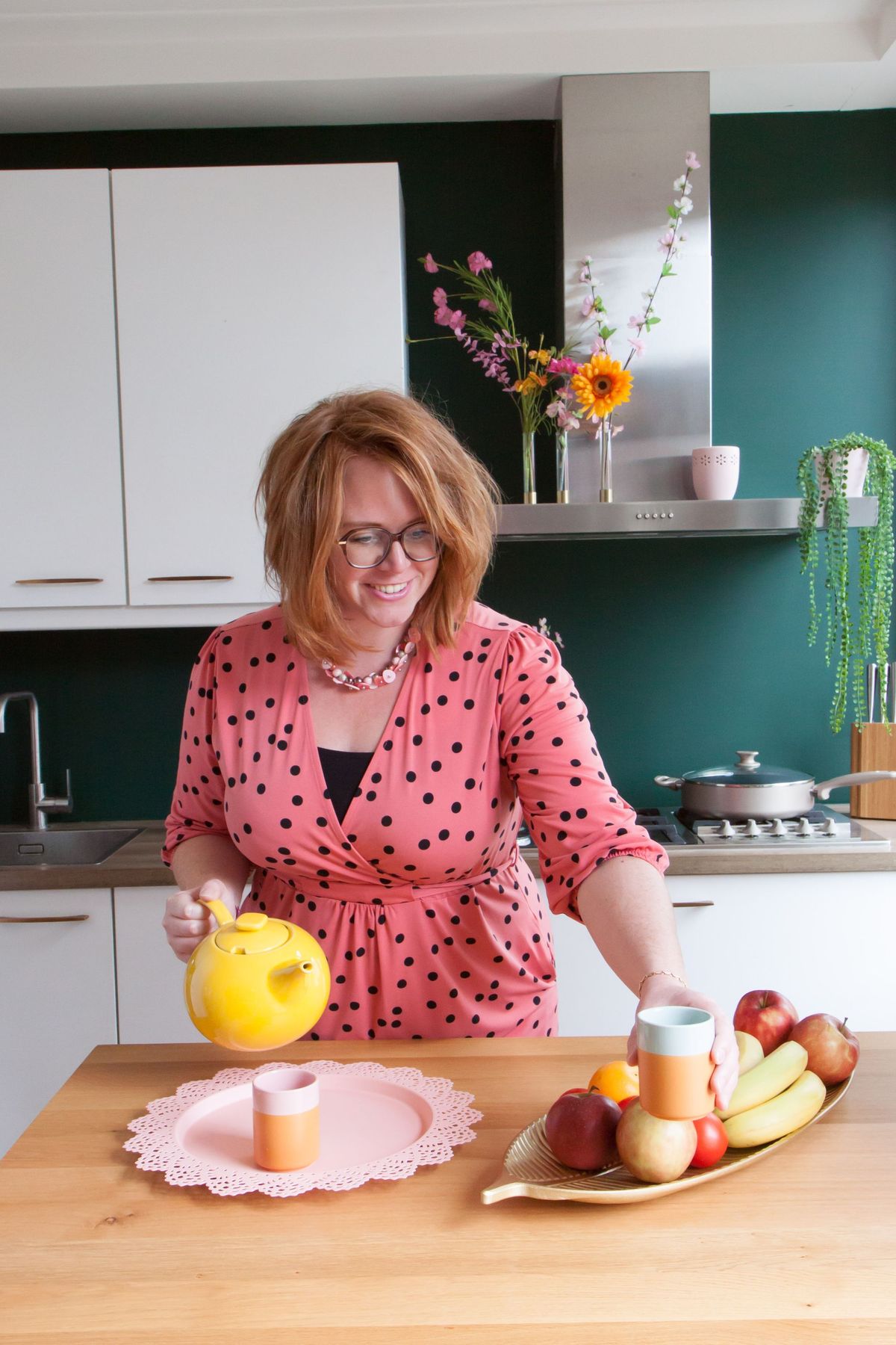 Roodharige vrouw met theepot in groene keuken, fruitschaal op bar